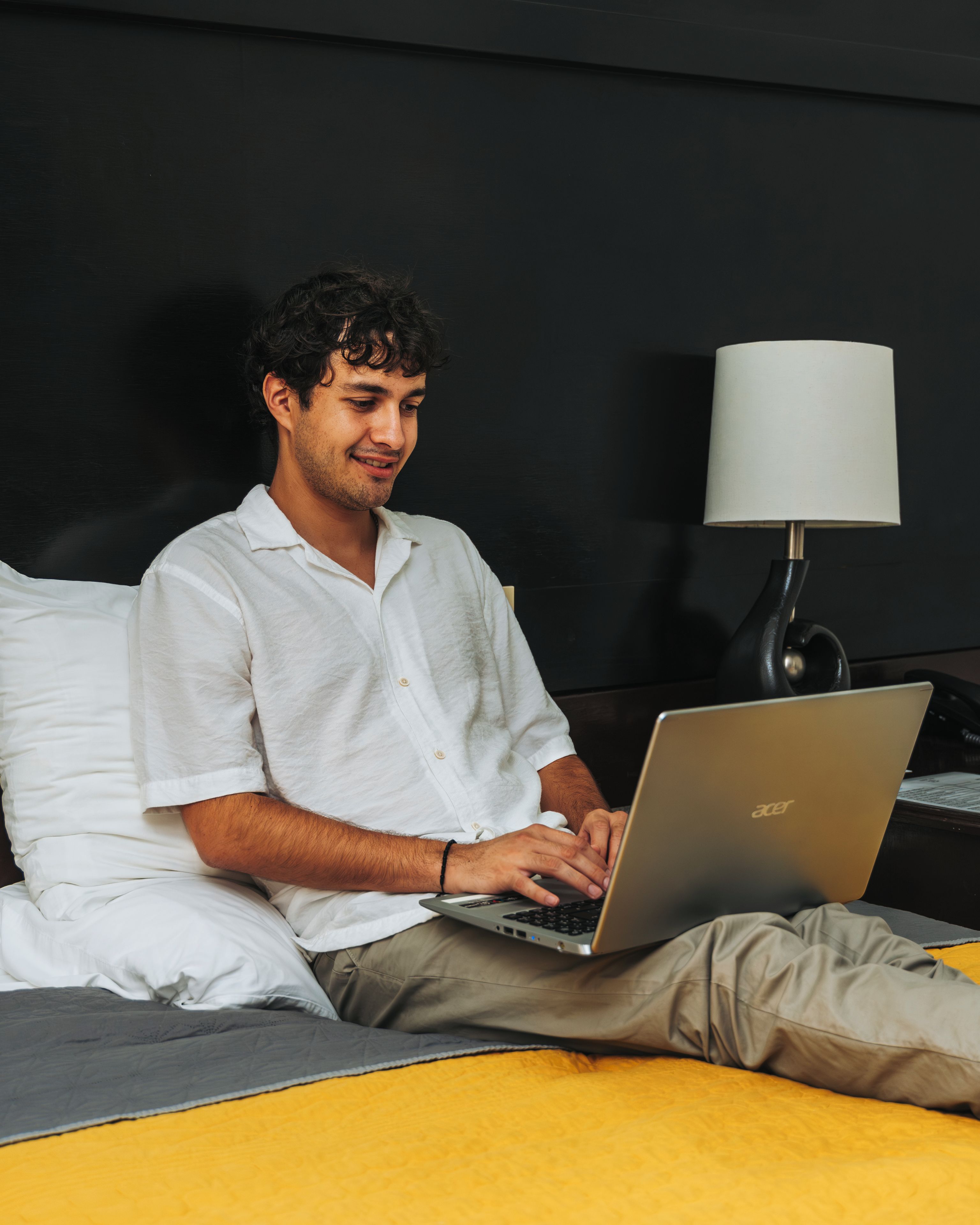 Eine Person sitzt auf einem Bett mit einem Laptop, einer Lampe und einem Kopfkissen in einem Zimmer mit einem dunklen Kopfteil.