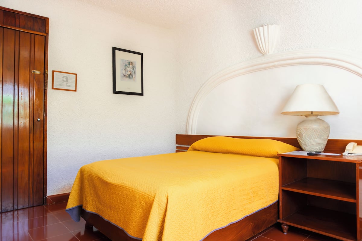 Una cama bien hecha con una colcha amarilla, una mesita de noche de madera con una lámpara y un cuadro enmarcado en la pared.