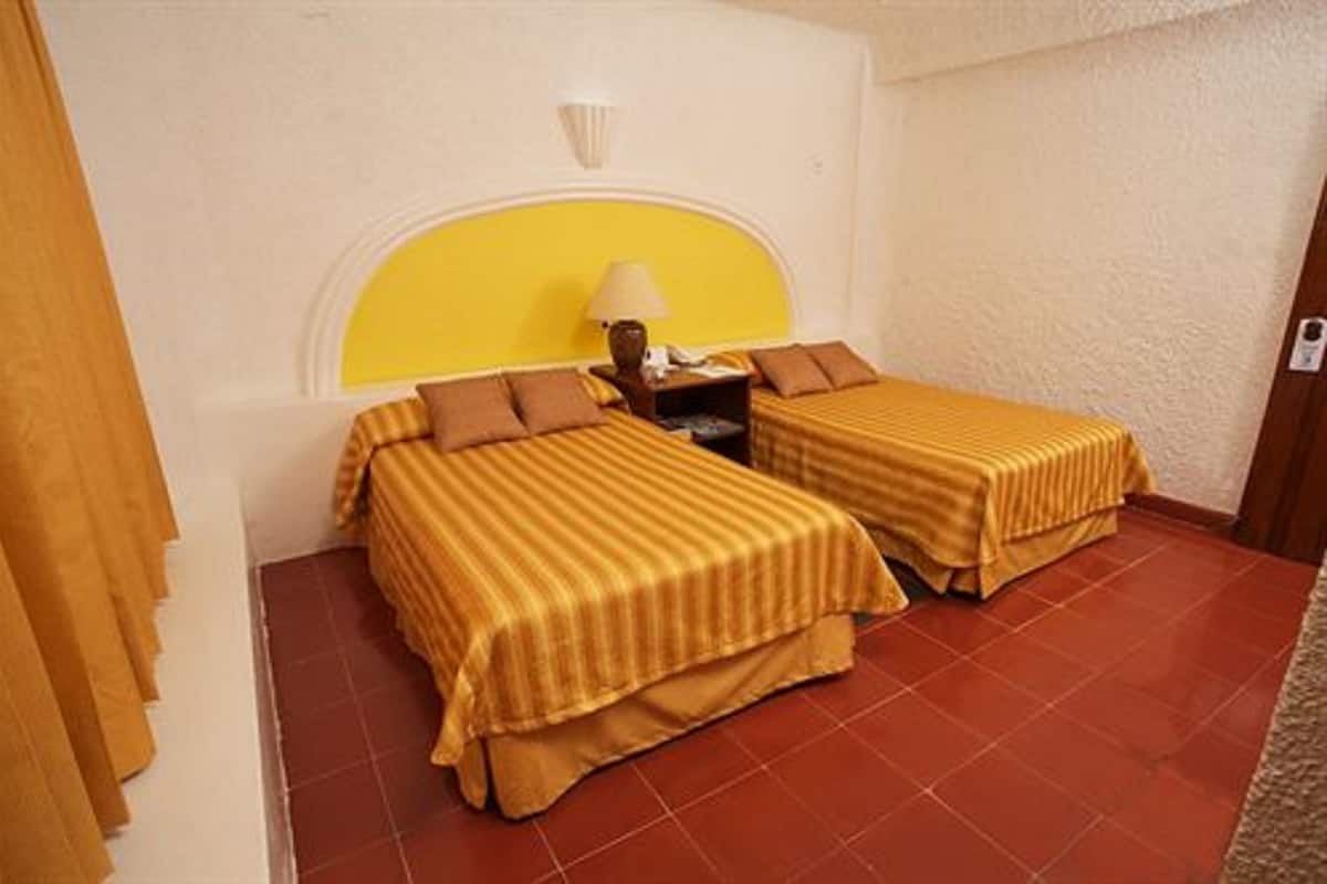 Habitación de hotel con dos camas, una mesita de noche, una lámpara y una pared de acento amarilla.