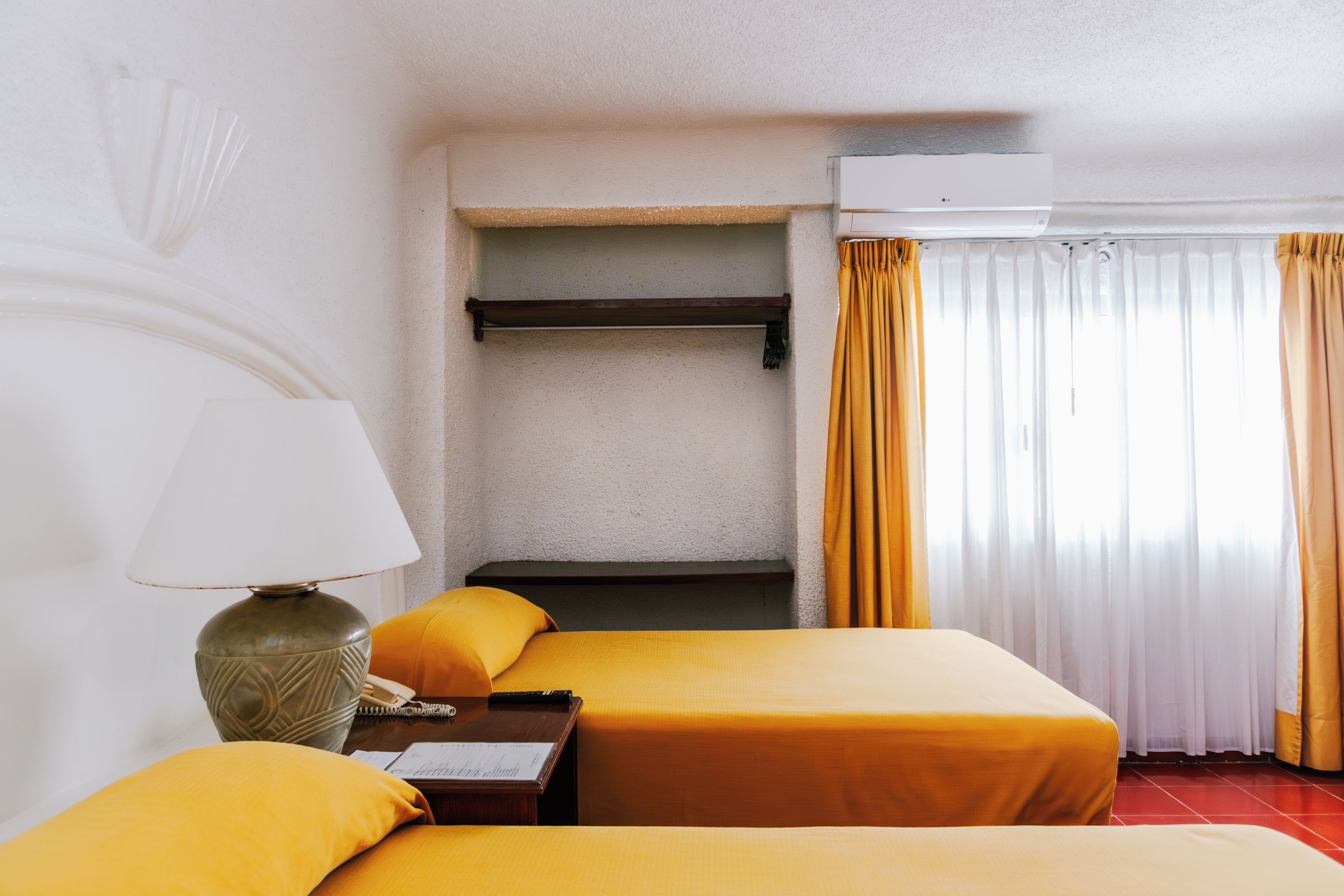 Une chambre d’hôtel avec deux lits, une table de chevet avec une lampe, une petite étagère et une fenêtre avec des rideaux.