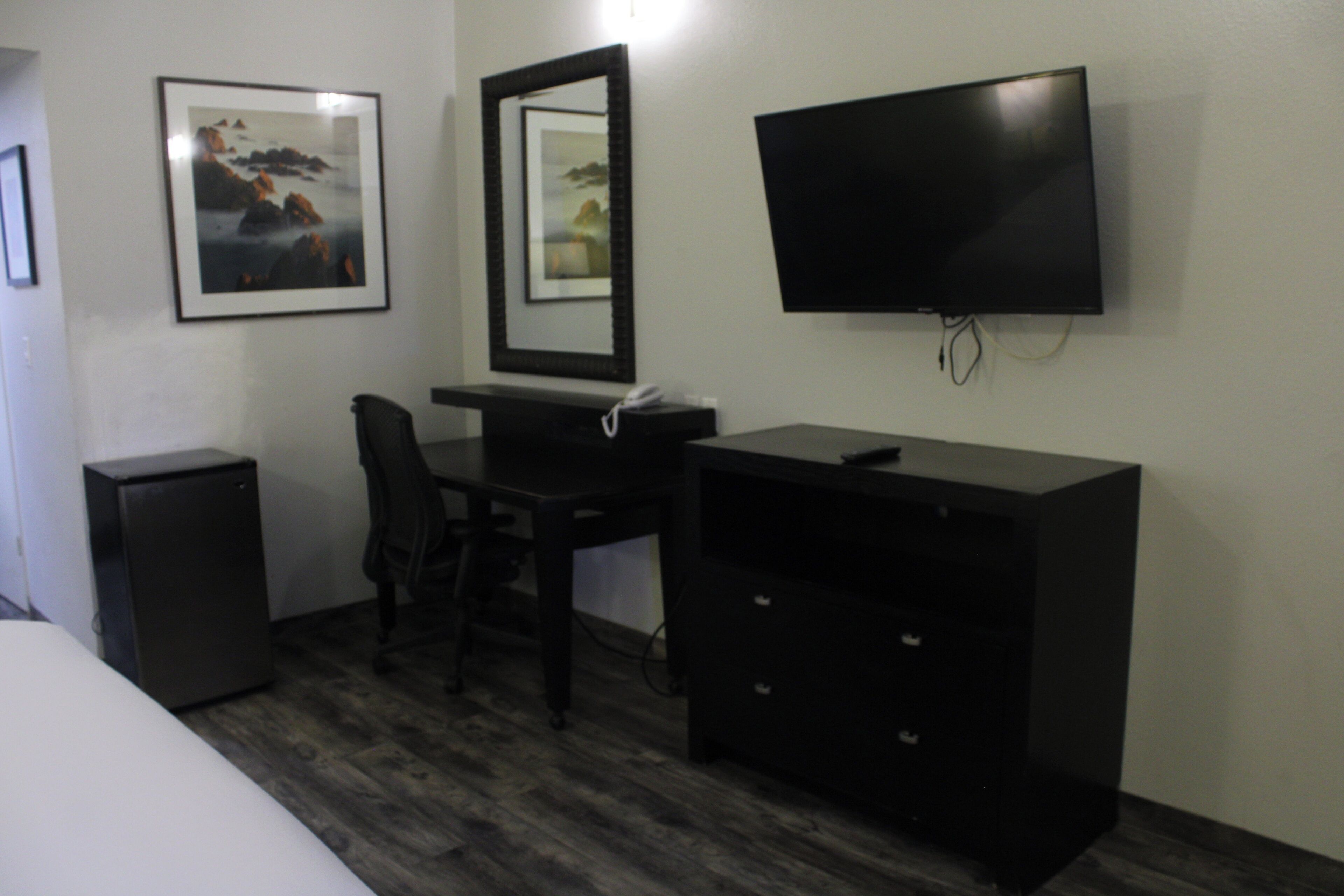 A hotel room with a bed, a desk with a chair, a TV, a small fridge, and two framed pictures on the wall.