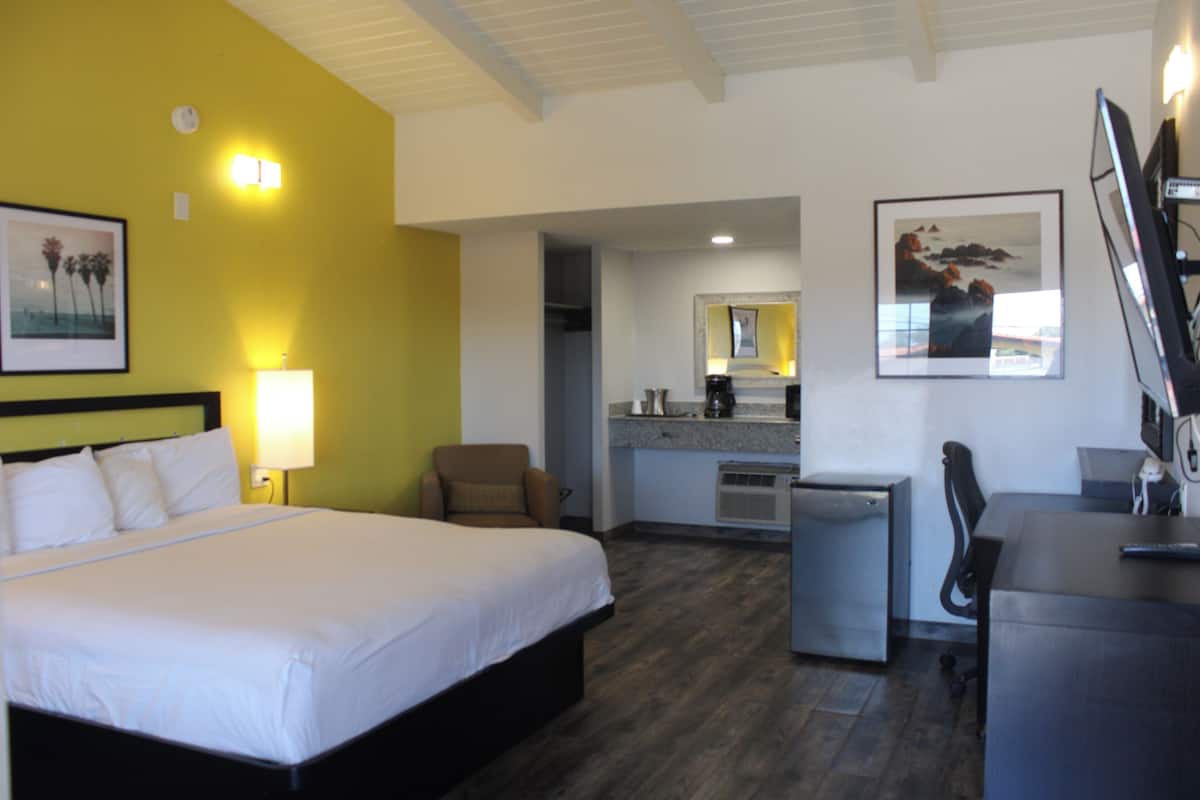 A hotel room with a bed, a desk, a chair, a kitchenette, and artwork on the walls.