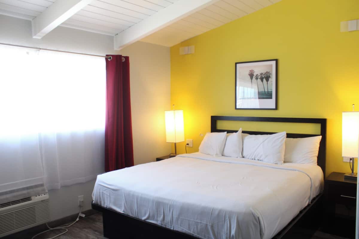 A hotel room with a bed, two bedside lamps, a heater, a window with curtains, and a picture on the wall.