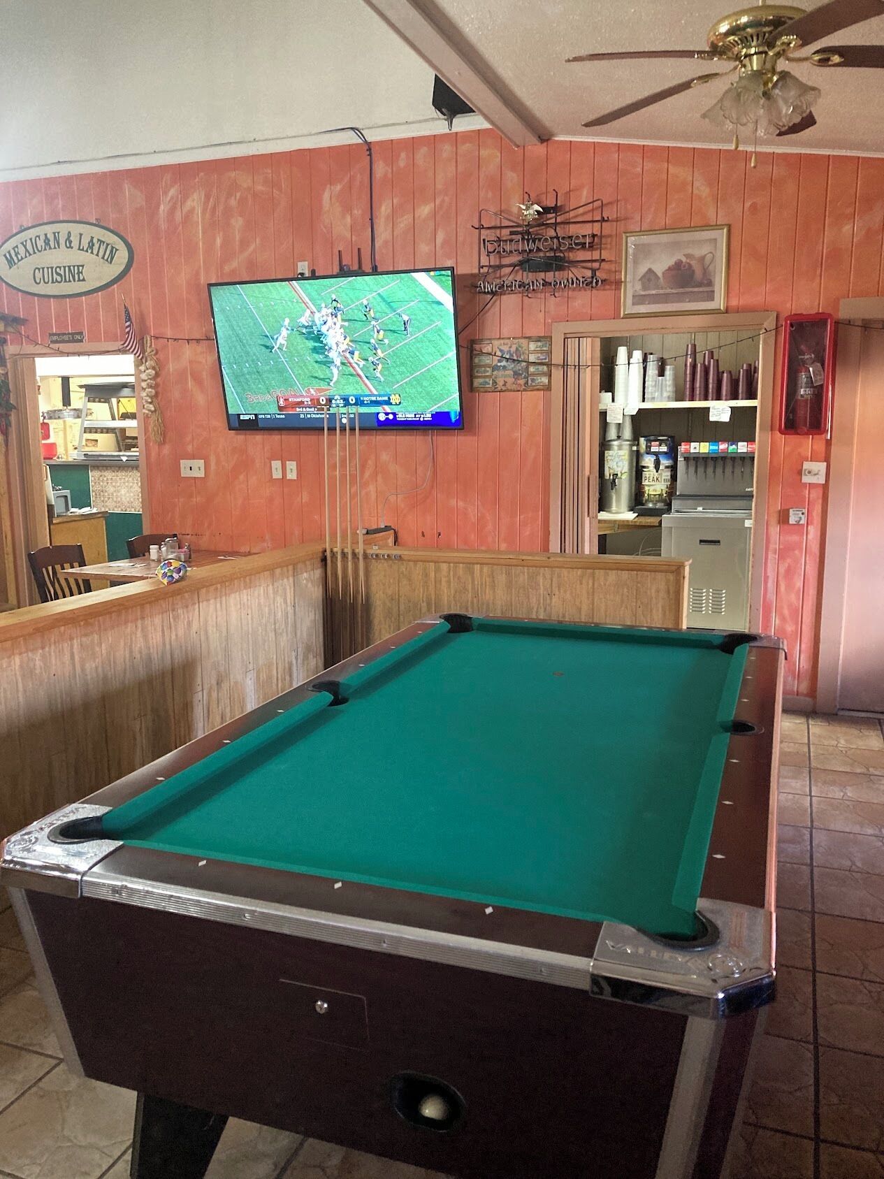Ein Billardtisch in einem Raum mit einem an der Wand befestigten Fernseher, der ein Sportspiel zeigt.
