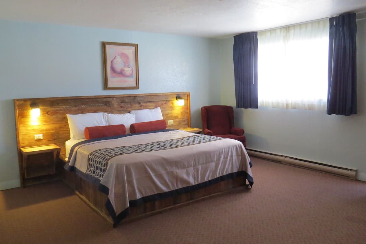 A bedroom with a wooden headboard, a bed with white and red bedding, a nightstand with a lamp, a red chair, and a window with curtains.