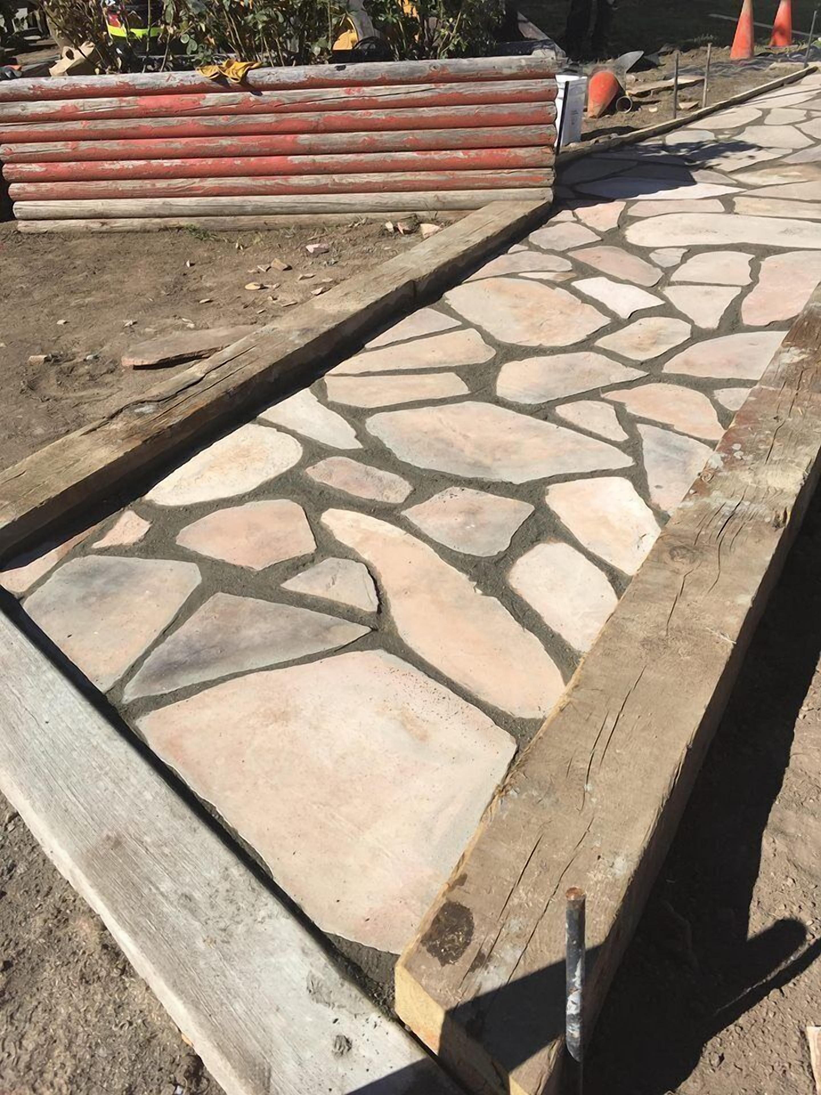 Un camino de piedra con bordes de madera que conduce a una obra en construcción con conos de tráfico naranjas.