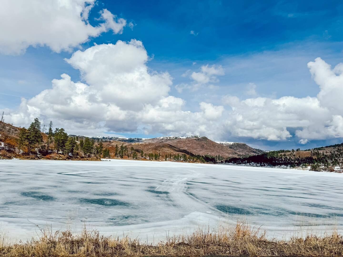 Ein zugefrorener See mit umliegenden Hügeln und vereinzelten Bäumen unter einem teils bewölkten Himmel.