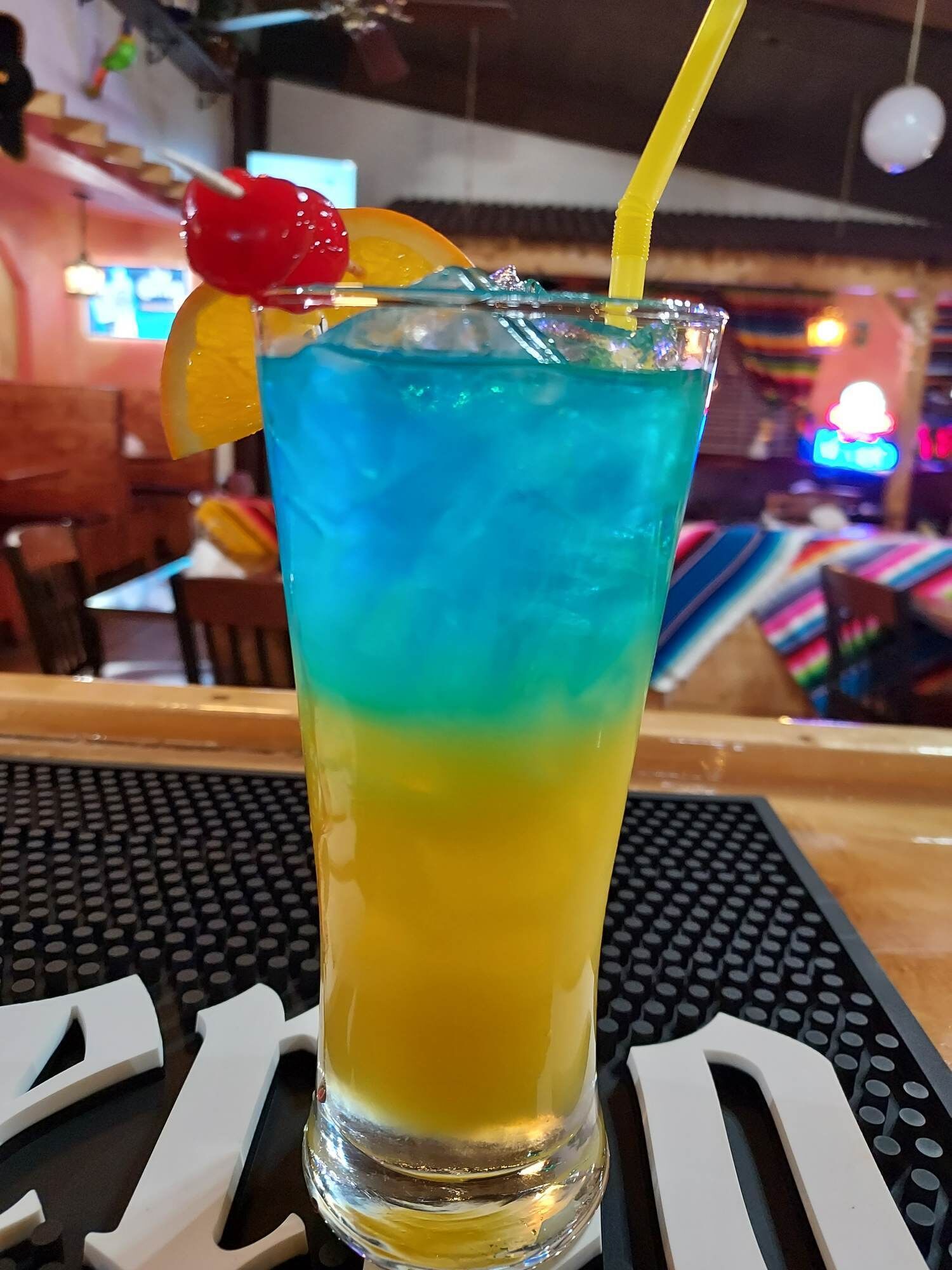 Un grand verre contenant un liquide dont la couleur dégrade du bleu au jaune, une paille jaune et une cerise rouge posée sur une tranche d’orange.