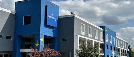 Ein modernes Hotel mit blauer Fassade, zahlreichen Fenstern und einem Parkplatz mit Autos.