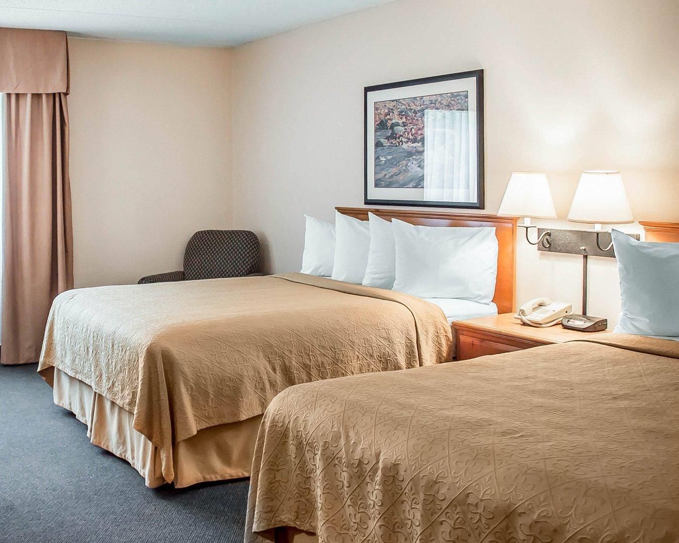 A hotel room with two beds, a chair, a desk with a lamp, and a painting on the wall.