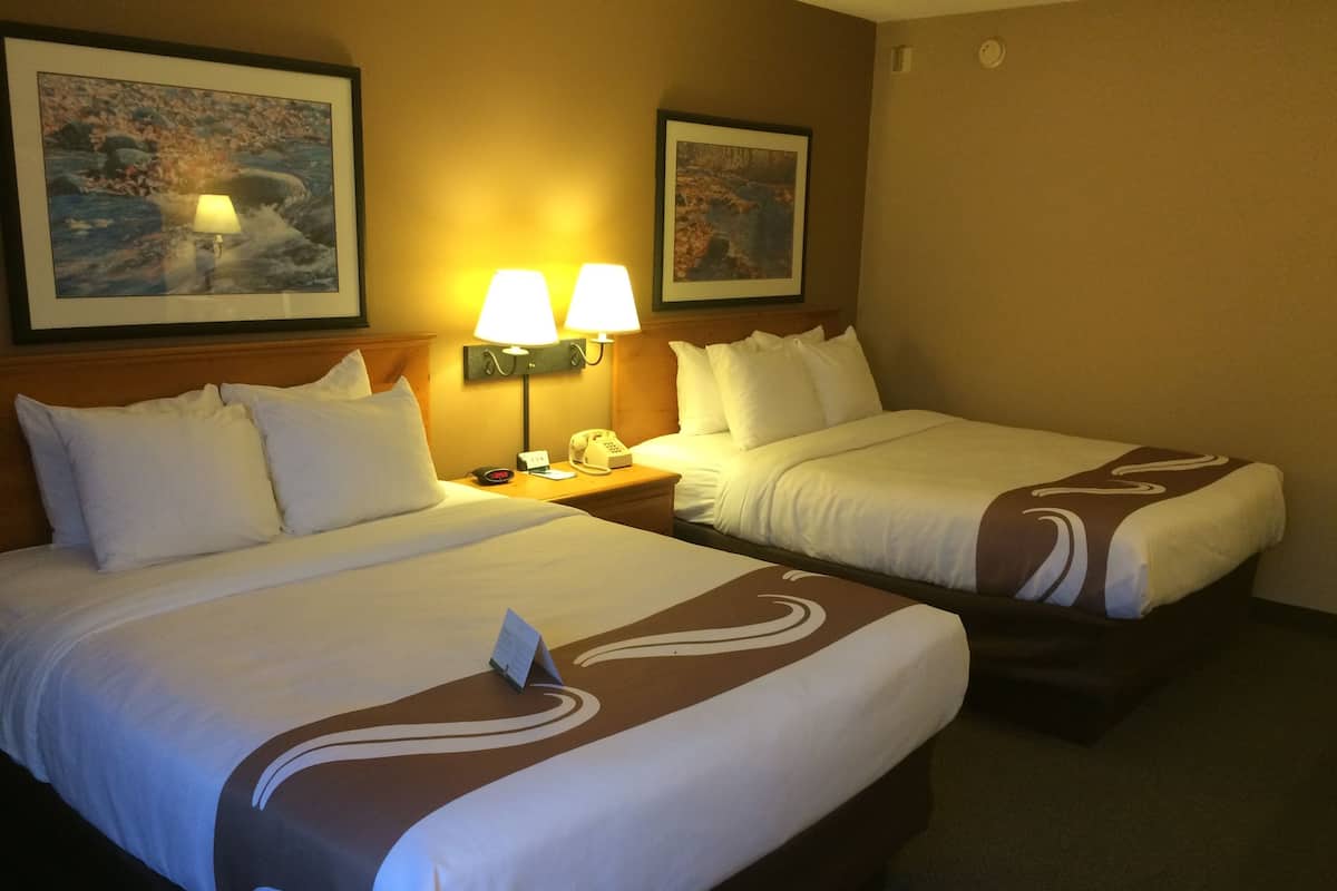 A hotel room with two beds, each with white bedding and brown decorative pillows. There are two wall-mounted lamps on the nightstands, and two framed pictures on the walls.