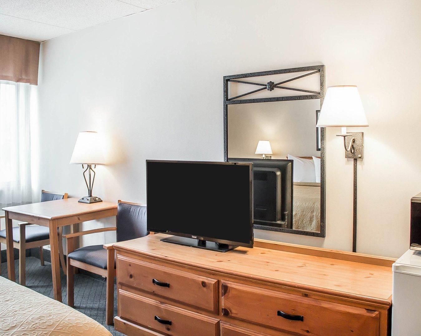 A hotel room with a TV on a wooden dresser, a bed with a patterned bedspread, a desk with a lamp, and a wall-mounted mirror.