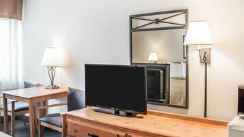 A hotel room with a TV on a wooden dresser, a bed with a patterned bedspread, a desk with a lamp, and a wall-mounted mirror.