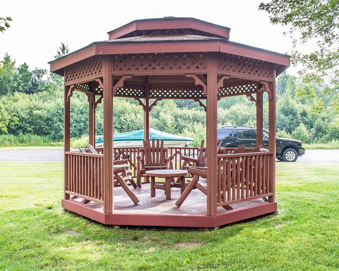 A wooden gazebo with a lattice roof, seating area, and a view of a parking lot and greenery.