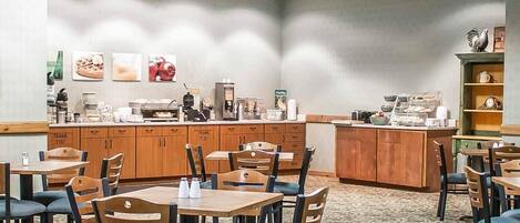 A hotel dining area with multiple tables and chairs, a counter with coffee machines, and a wall with framed pictures.