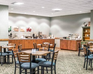 A hotel dining area with multiple tables and chairs, a counter with coffee machines, and a wall with framed pictures.