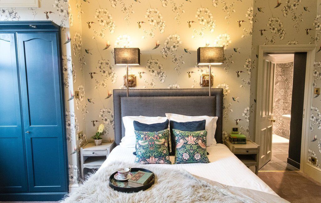 A bedroom with a large bed, floral wallpaper, two bedside lamps, a nightstand, and a wardrobe.