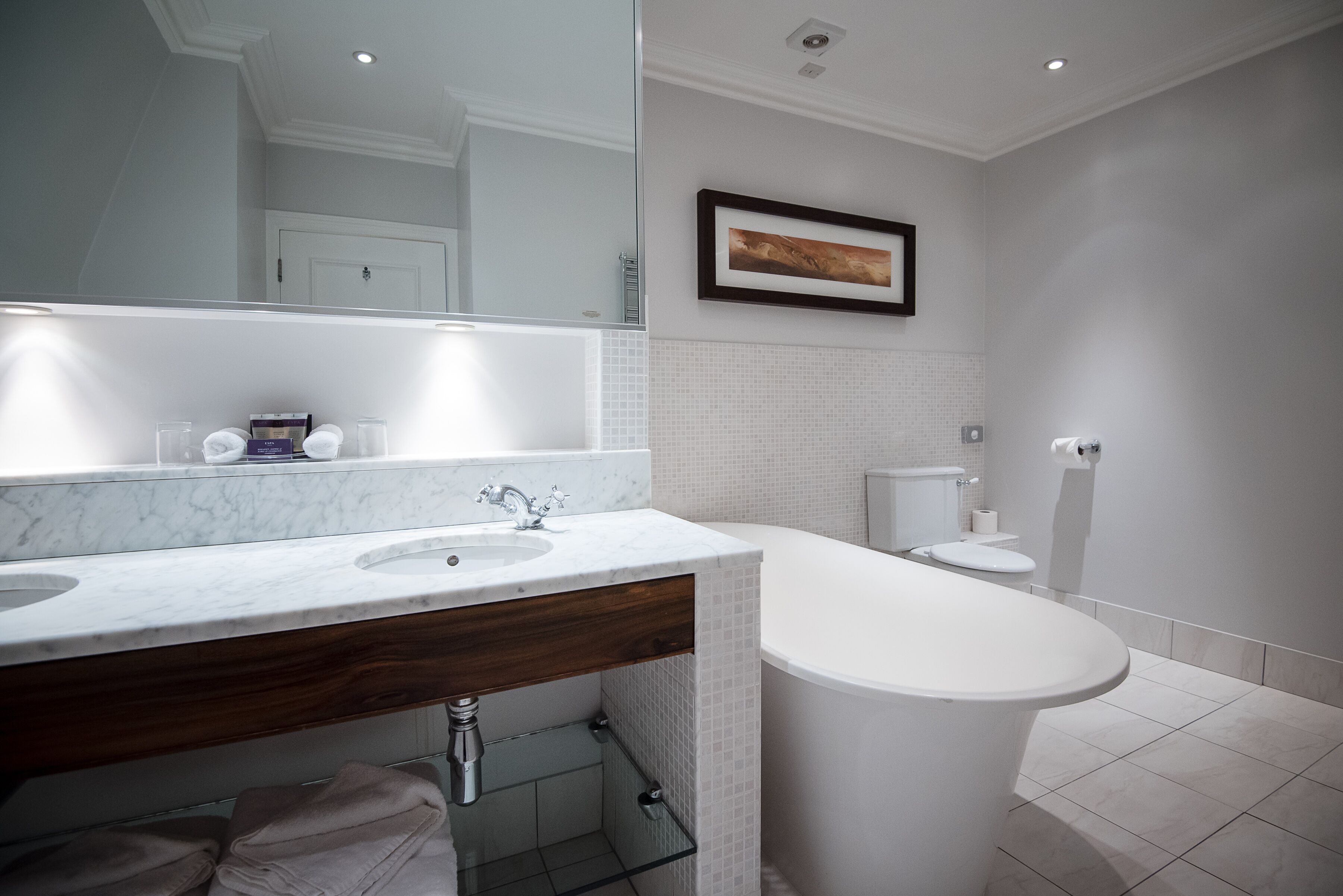 A modern bathroom with a marble sink, wooden vanity, a large mirror, a framed picture on the wall, and a standalone bathtub.