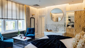 A modern hotel room with a large bed, a sitting area with a blue armchair, a round mirror, and a bathroom with a white bathtub and marble walls. - The Spa Hotel (Royal Tunbridge Wells)