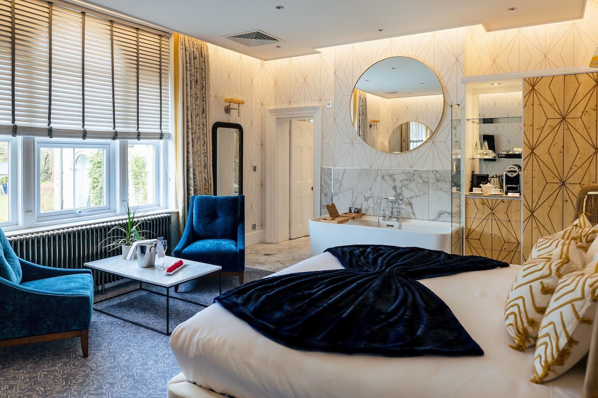 Una habitación de hotel moderna con una cama grande, una zona de estar con un sillón azul, un espejo redondo y un baño con una bañera blanca y paredes de mármol.
