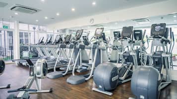 A gym with various cardio machines, wooden flooring, and large windows.