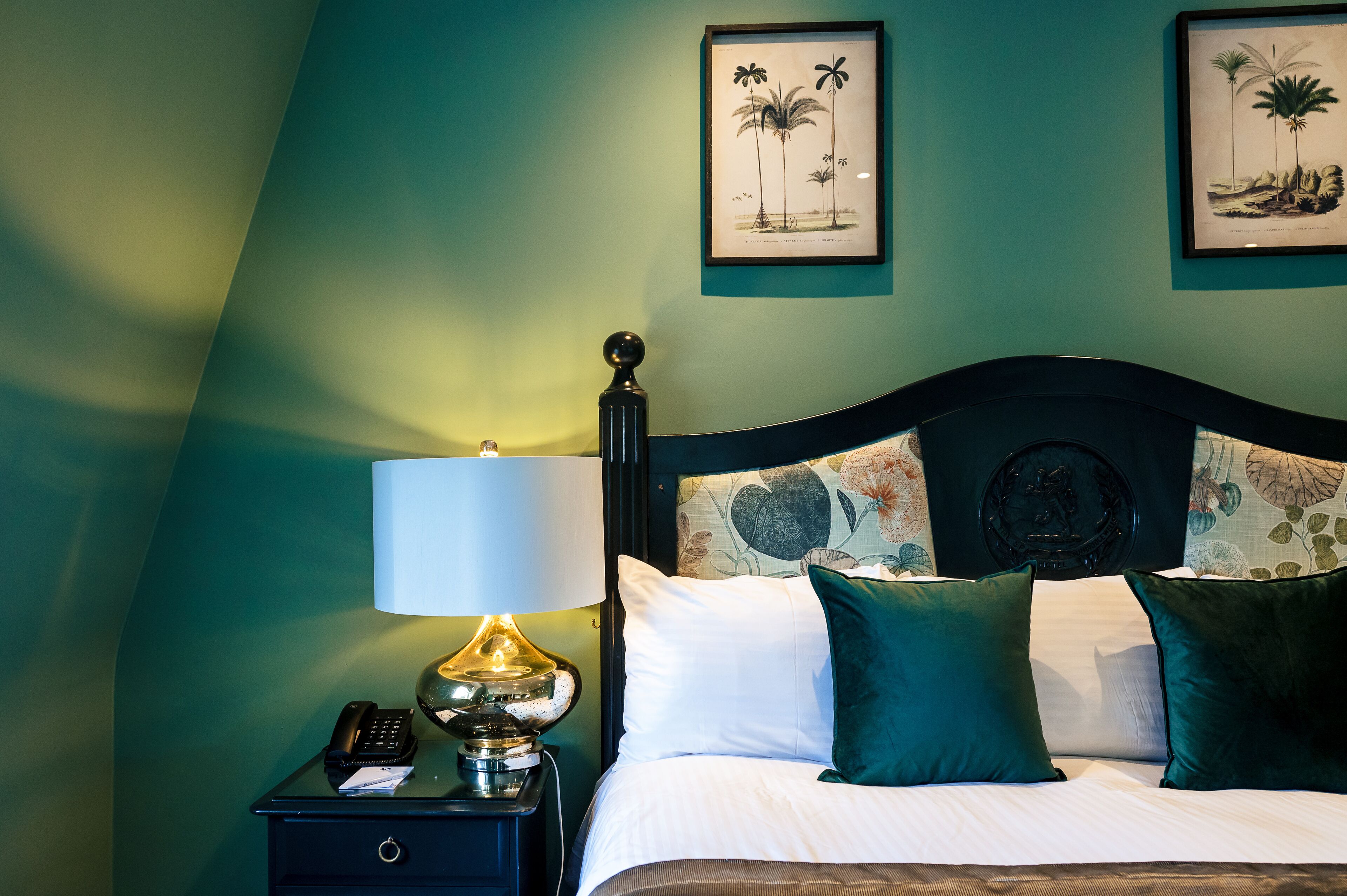 A neatly made bed with green and white pillows, a bedside lamp, and a nightstand with a phone and a lamp.