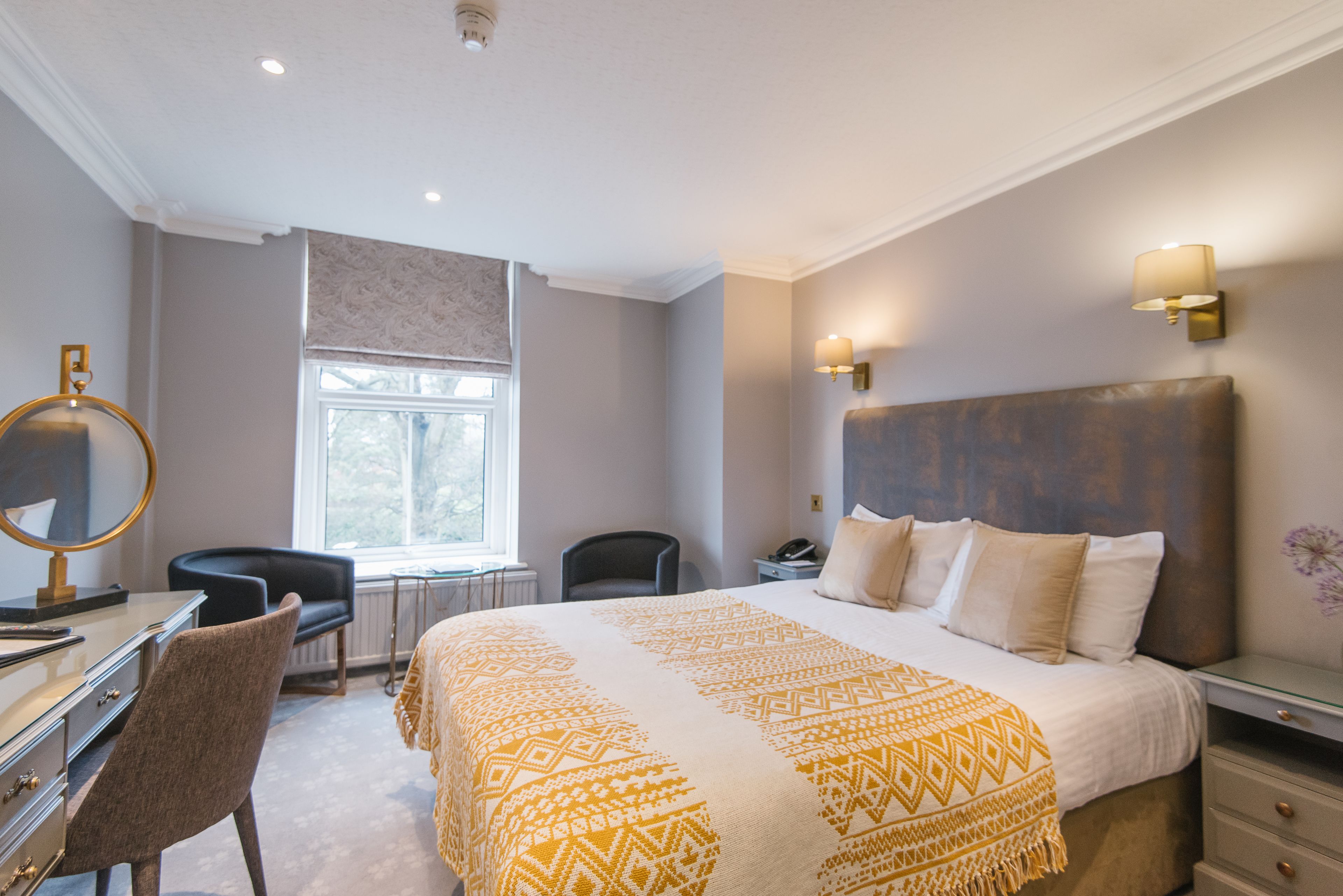 A hotel room with a large bed, a desk with a chair, a window, and a wall-mounted lamp.