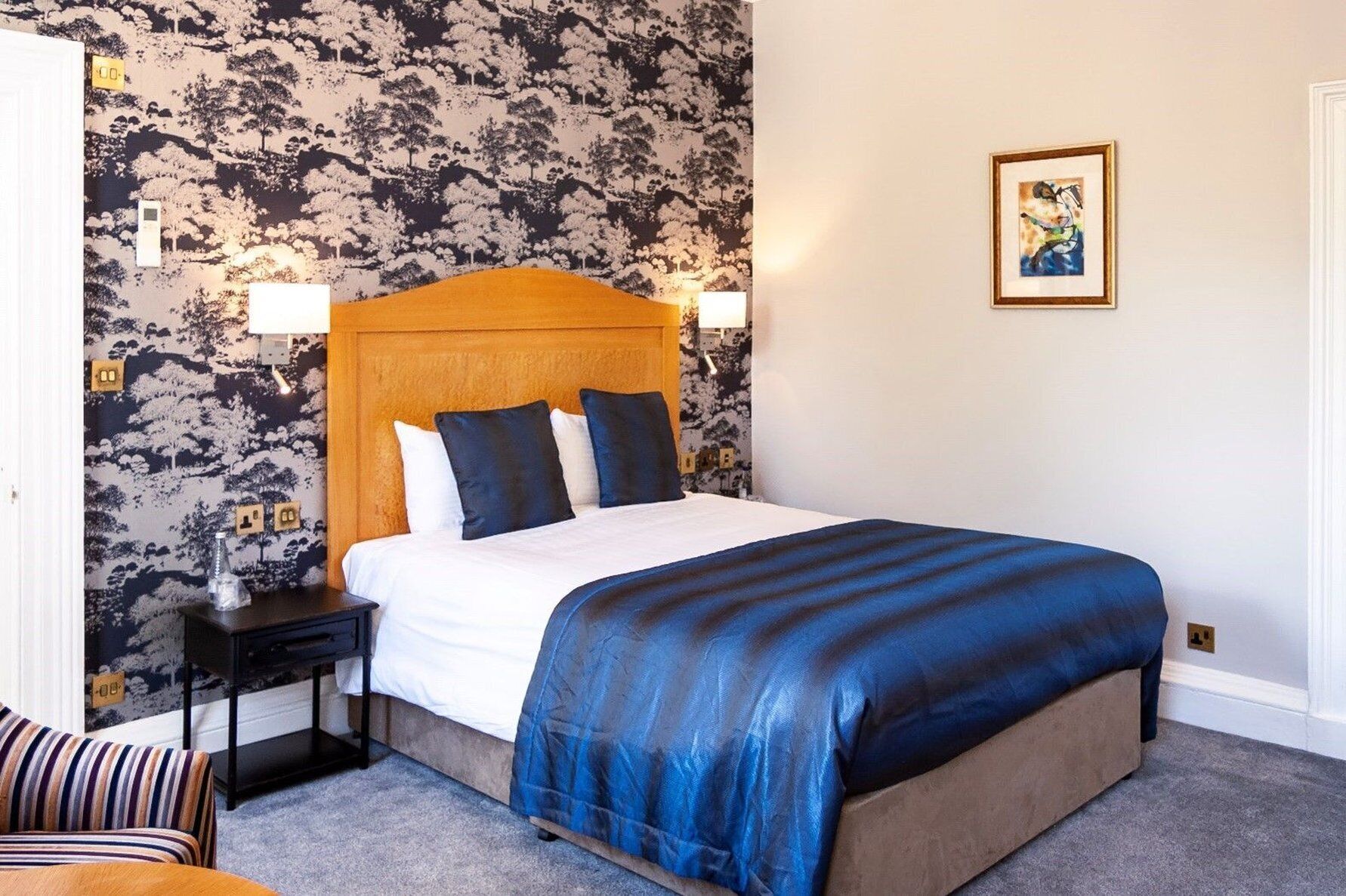 A hotel room with a bed, a nightstand, a lamp, a chair, and a wall with a patterned wallpaper.
