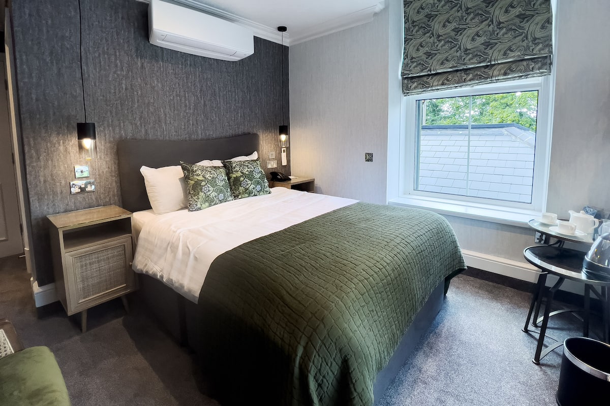 Una habitación de hotel moderna con una cama grande, mesitas de noche, una pequeña mesa con tazas, una ventana con vistas y un equipo de aire acondicionado.