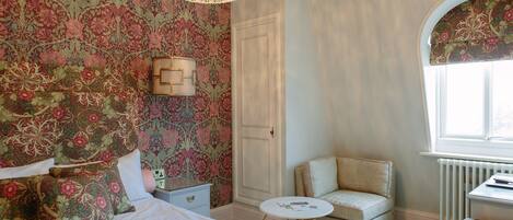 Un dormitorio con papel tapiz floral, una cama con muchos cojines, una mesita de noche, una silla, una pequeña mesa y una ventana con arco decorativo.