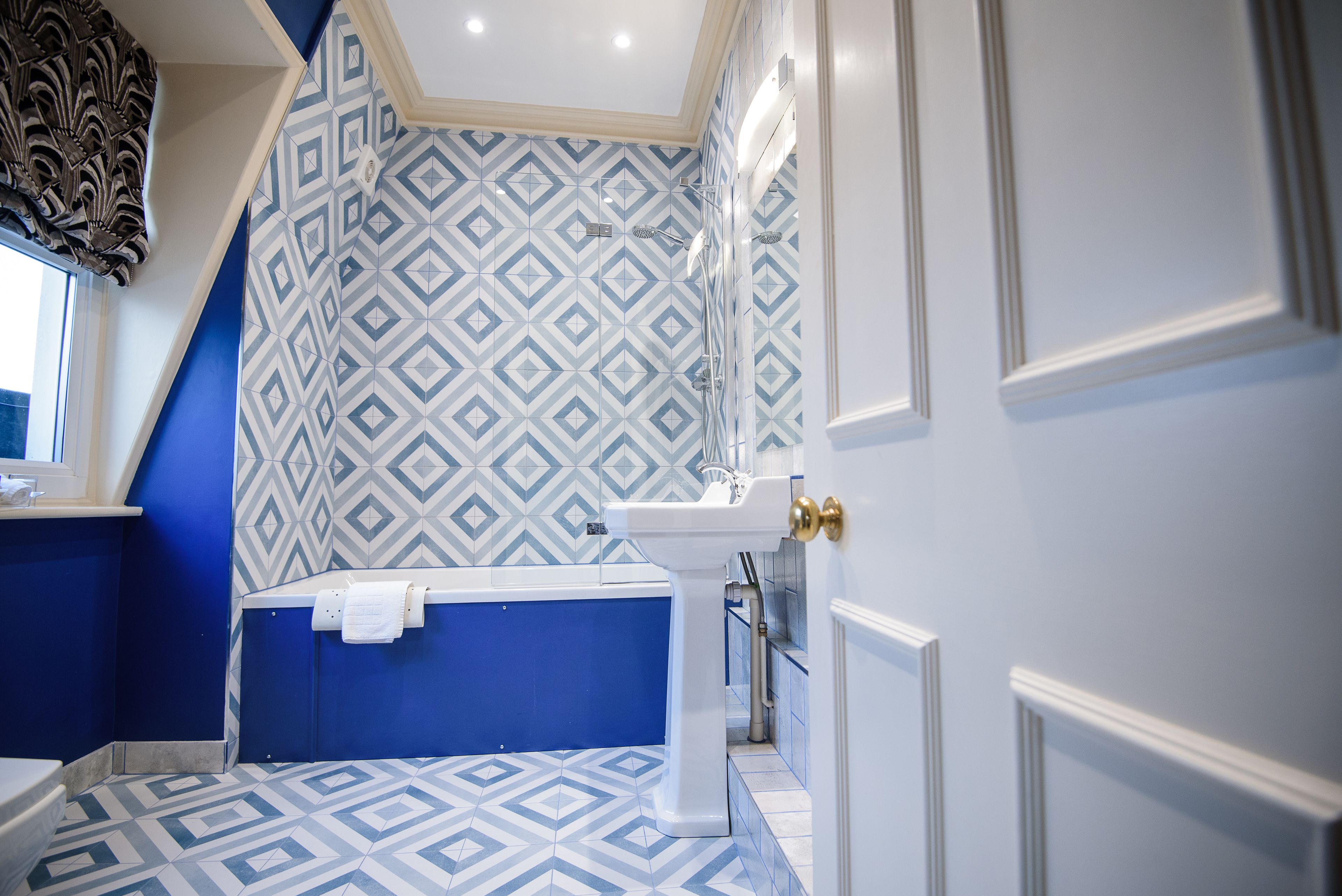 A bathroom with a geometric patterned tile floor, a white pedestal sink, and a bathtub with a blue wall.
