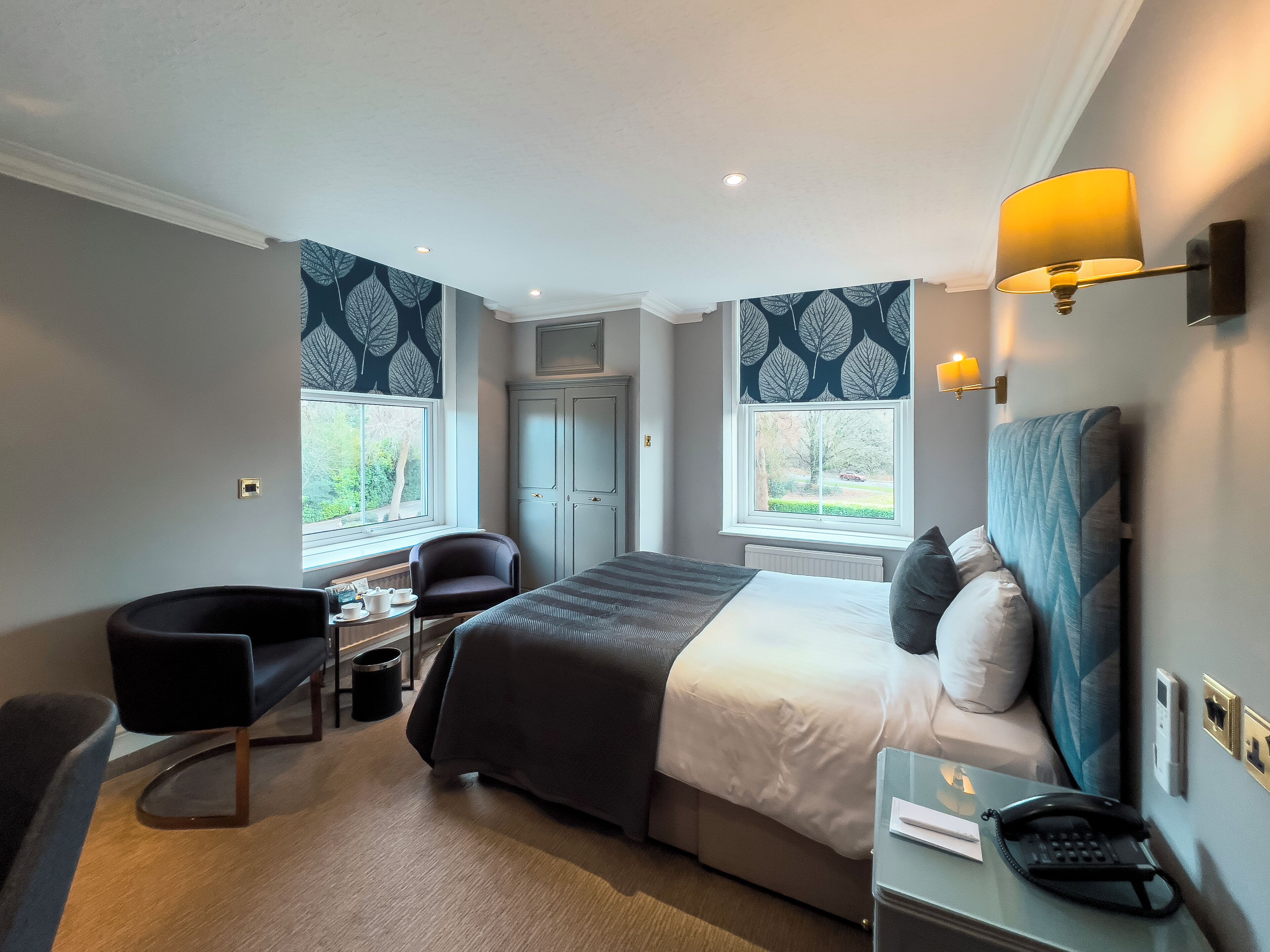 A hotel room with a bed, bedside table, lamp, chair, and a view of the outdoors through two windows.