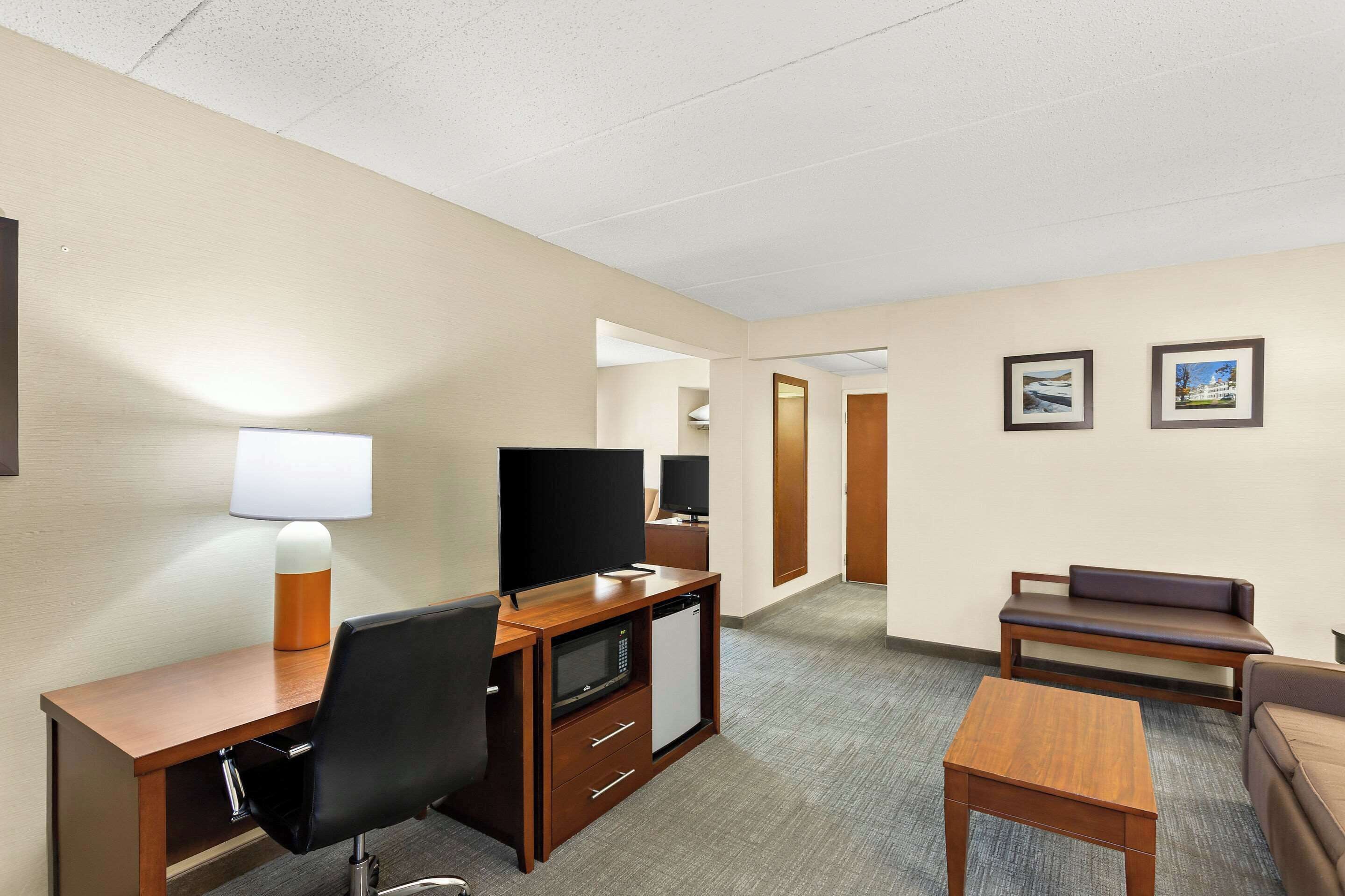 A hotel room with a wooden desk, a chair, a television, a microwave, a refrigerator, a sofa, and two framed pictures on the wall.