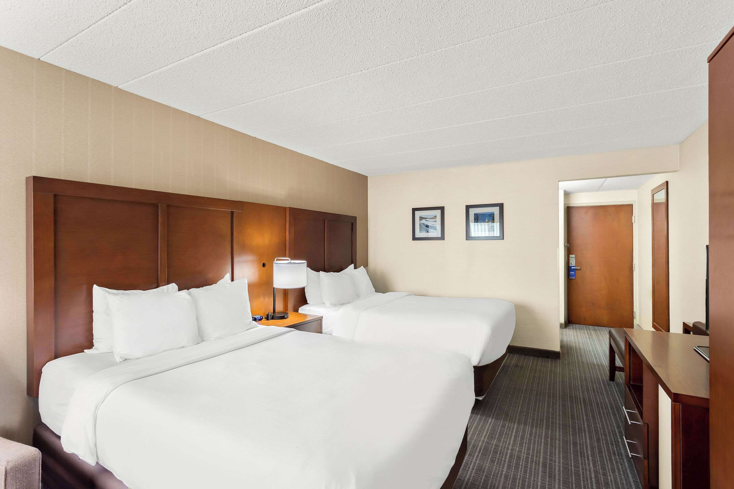 Habitación de hotel con dos camas, un escritorio, una lámpara y una puerta que da a otra habitación.