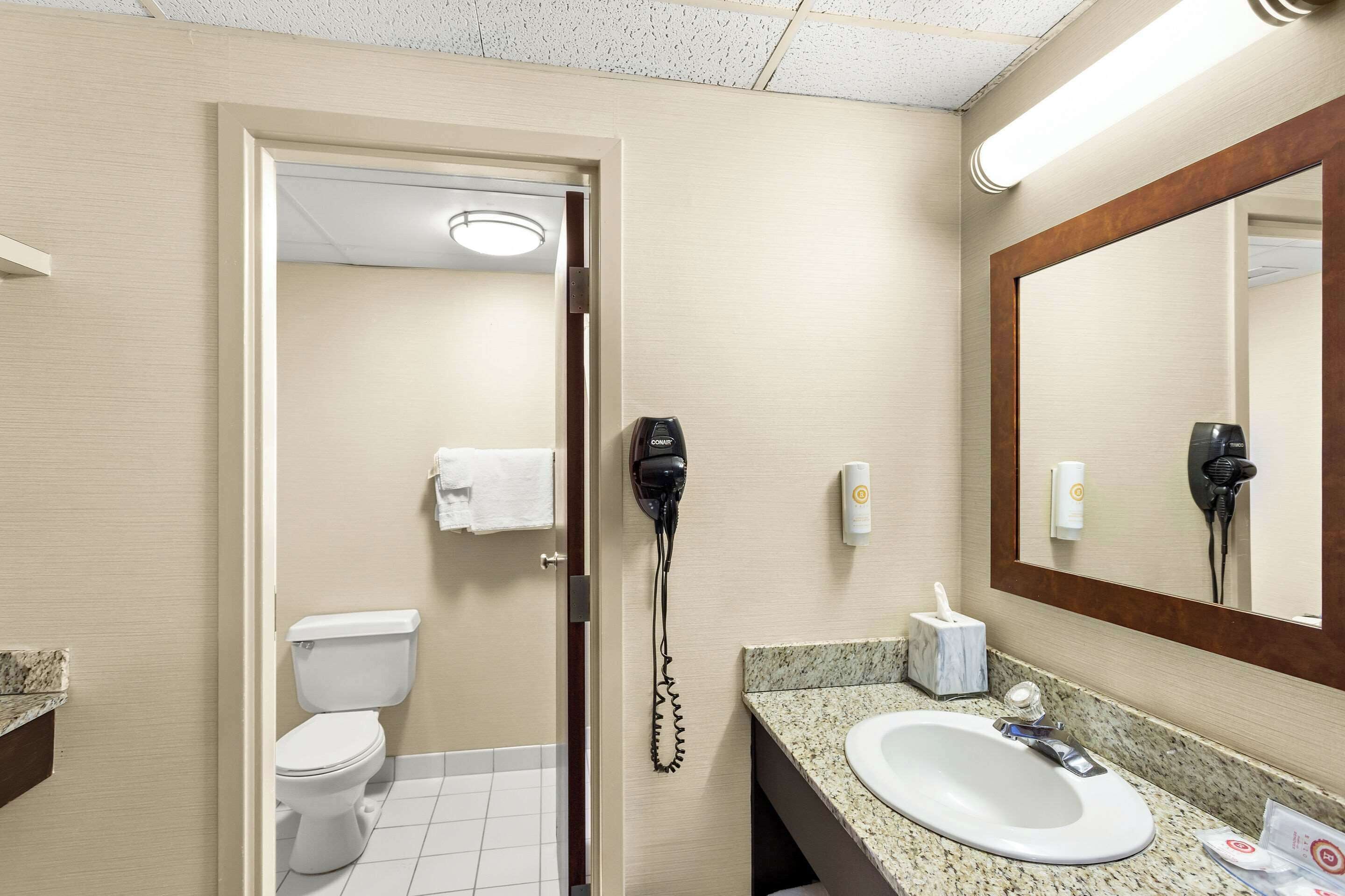 Un baño de hotel con inodoro, lavamanos, secador de pelo y espejo.
