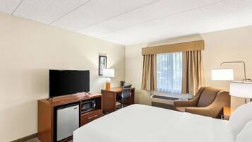 A hotel room with a bed, a desk with a chair, a television, a mini-fridge, and a window with curtains.