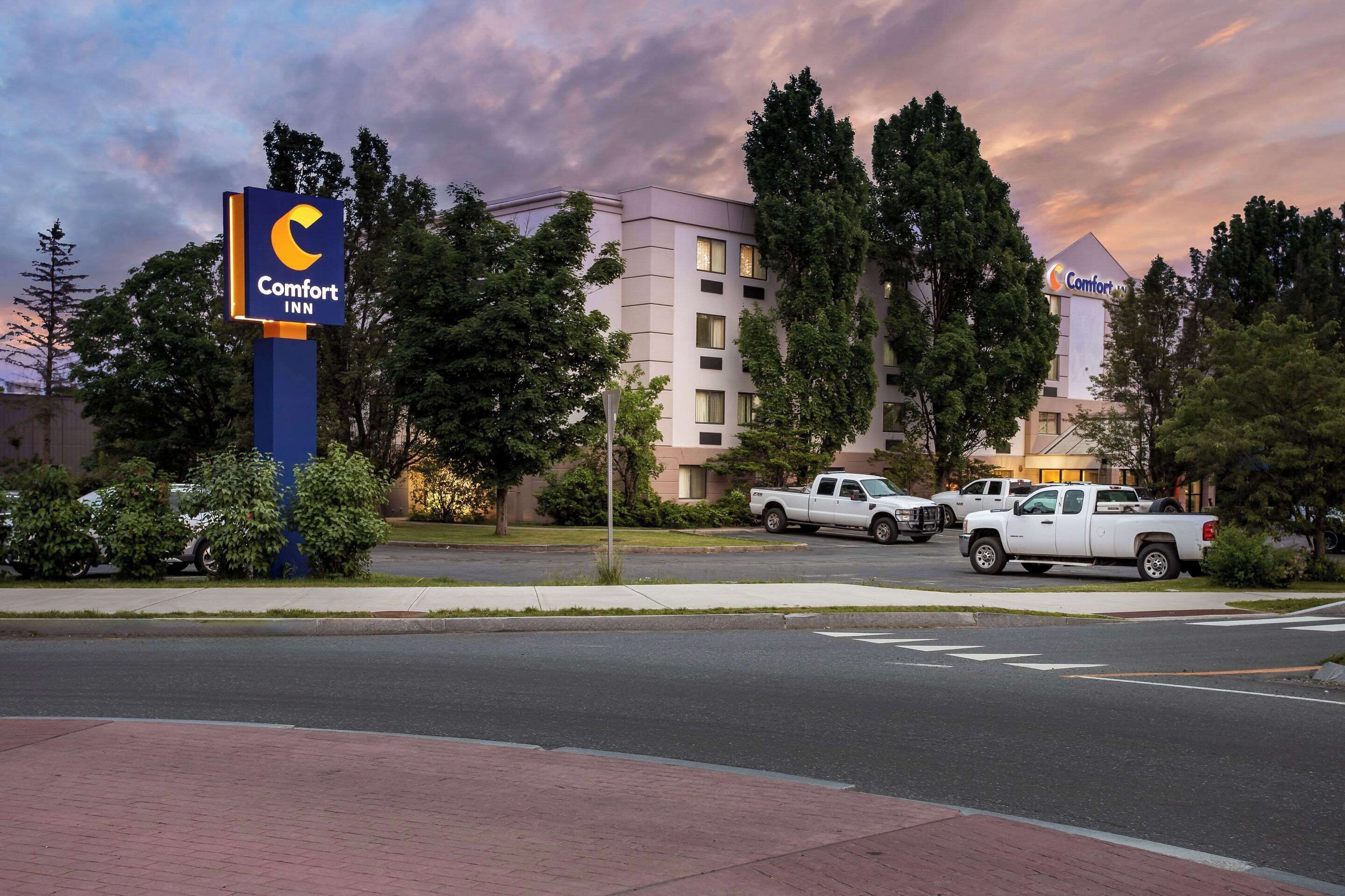 A Comfort Inn hotel with a prominent sign, a parking area with white pickup trucks, and a backdrop of trees and a cloudy sky.
