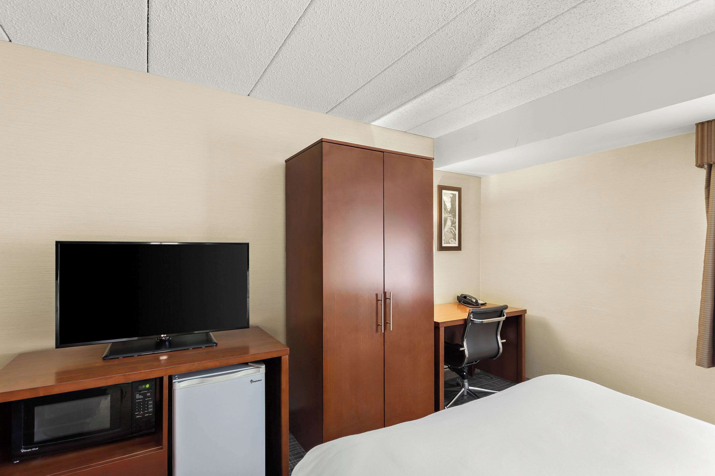 A hotel room with a bed, a television, a wooden wardrobe, a desk with a chair, and a microwave.