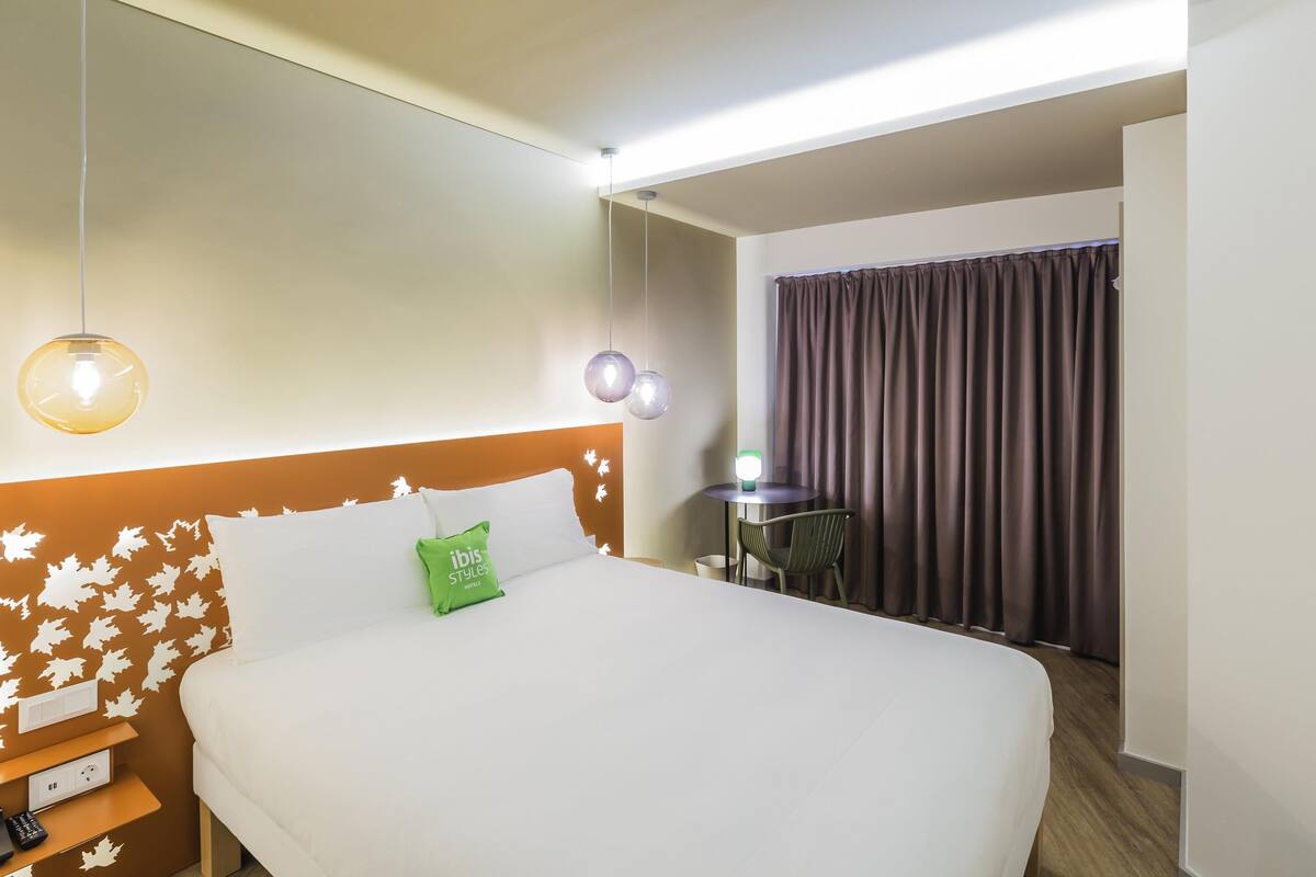 A hotel room with a bed, bedside table, lamp, and a view of a curtain and a small table.