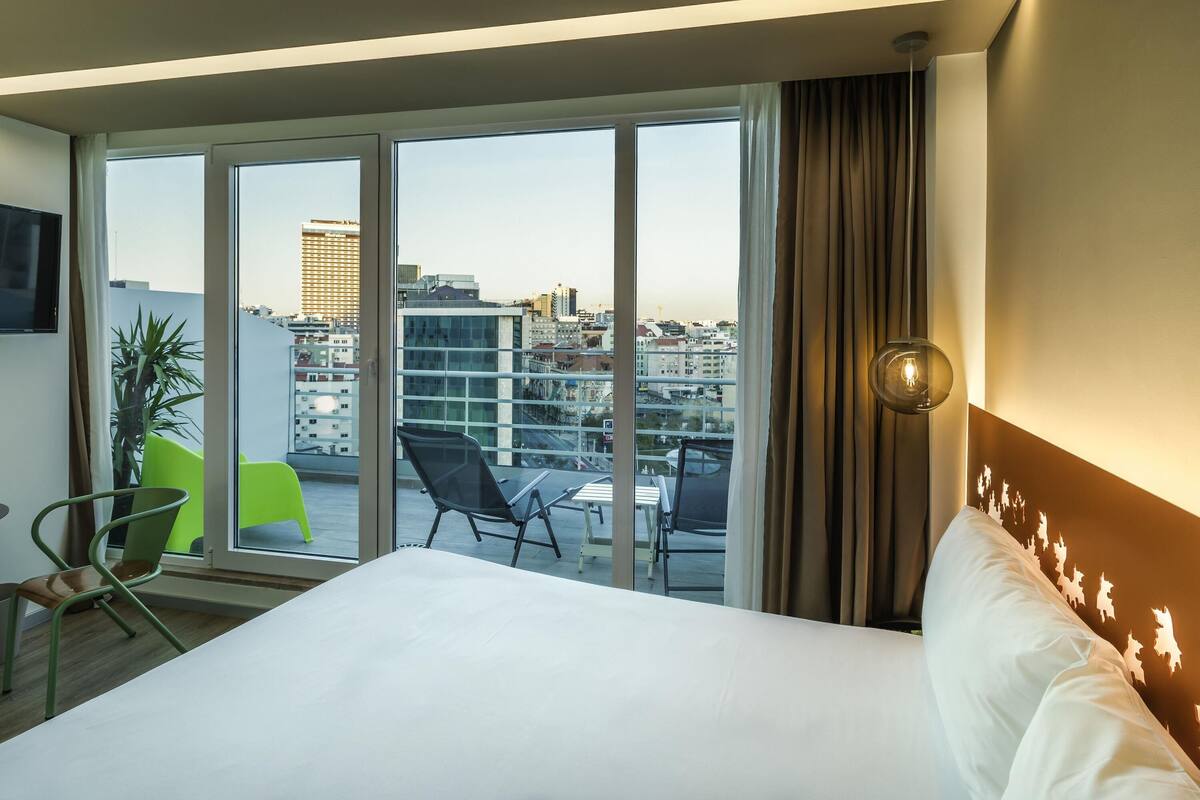 A hotel room with a large bed, a view of the cityscape, a TV mounted on the wall, and a plant.
