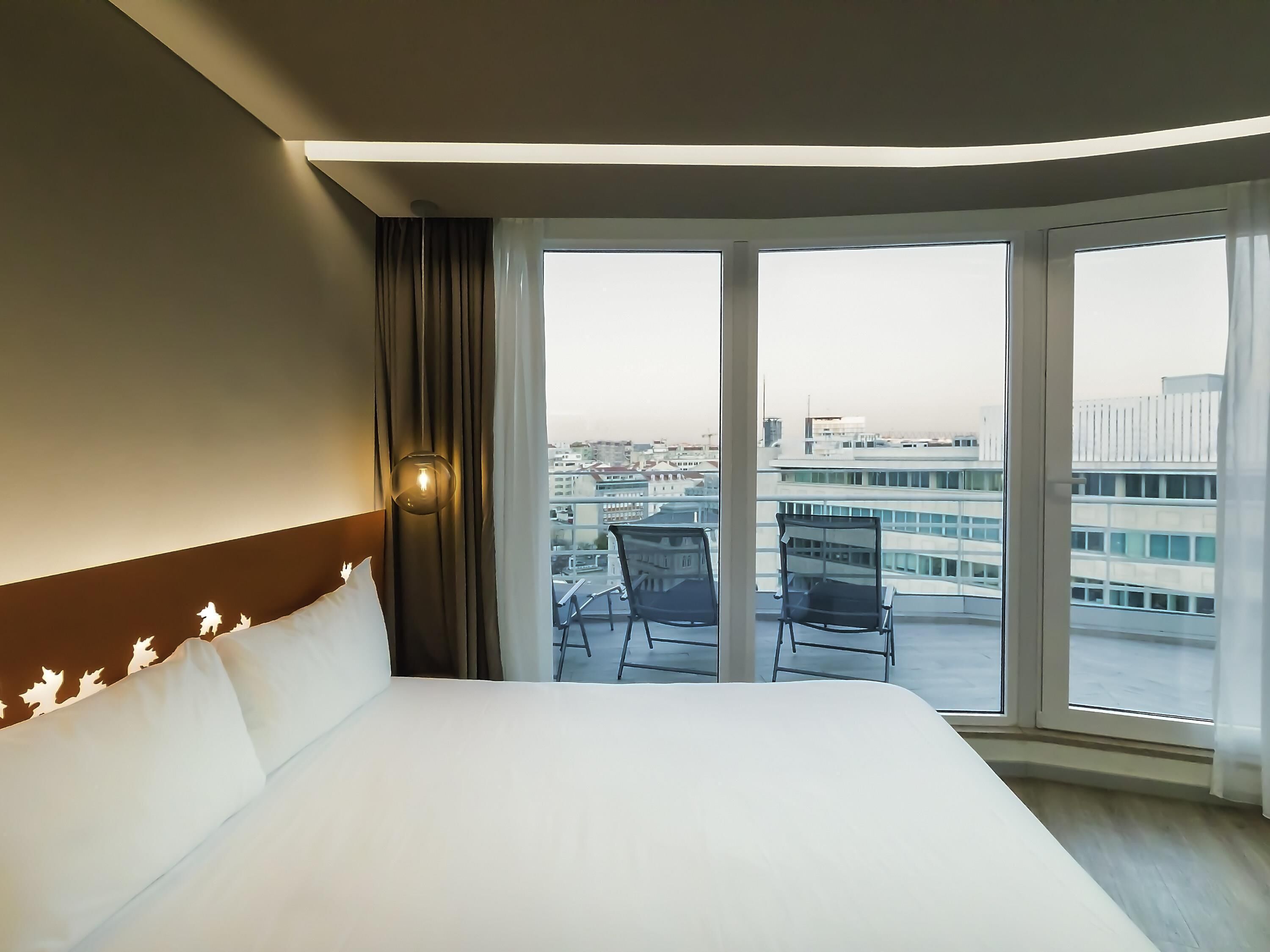 Habitación de hotel con una cama grande, vistas de la ciudad a través de amplios ventanales y un balcón con sillas.