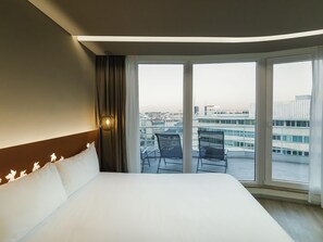 Ein Hotelzimmer mit einem großen Bett, Blick auf die Stadt durch große Fenster und einem Balkon mit Sitzgelegenheiten.