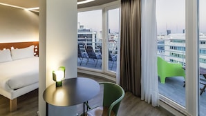 A hotel room with a bed, a round table, a green chair, and a balcony with a view of the city.