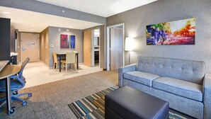 Ein modernes Hotelzimmer mit einer grauen Couch, einem kleinen Tisch, einem Schreibtisch mit Stuhl und einem farbenfrohen, abstrakten Gemälde an der Wand.