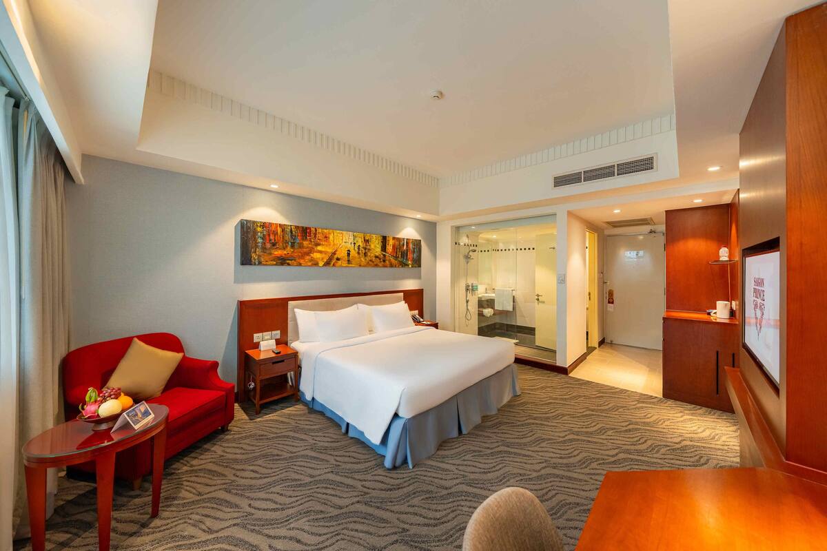 A hotel room with a bed, a red sofa, a small table with fruit, a painting on the wall, and a shower area.
