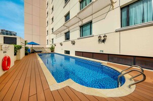 A pool area with a tiled deck, a lifebuoy, and a building with windows and a balcony.