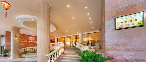 A hotel lobby with a marble reception desk, a staircase with a balustrade, and a sign on the wall.