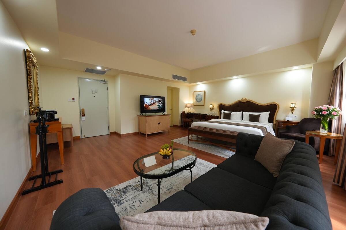 A hotel room with a large bed, a sofa, a TV, a desk, and a small table with fruit.
