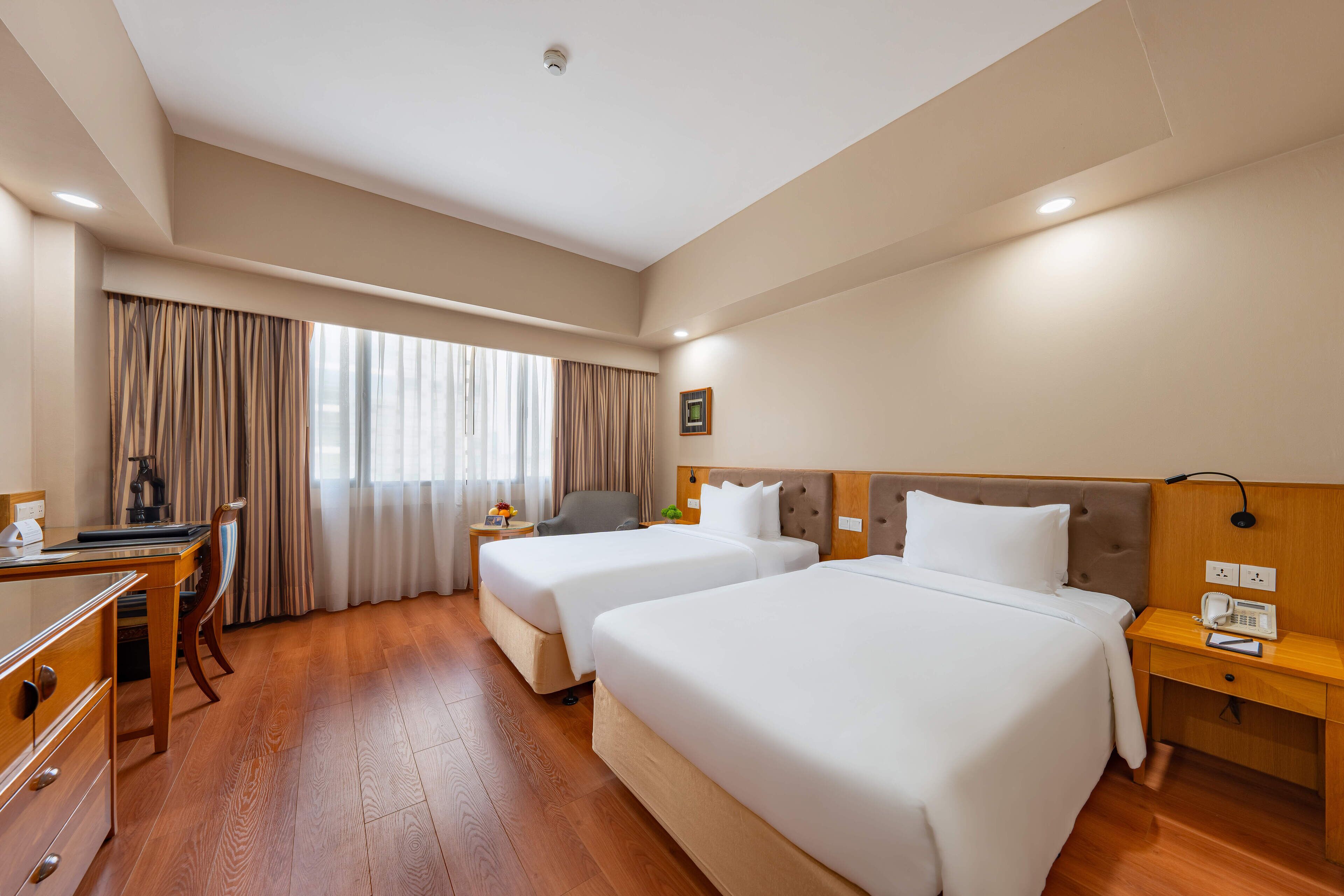 Une chambre d’hôtel avec deux lits, un bureau, une chaise, une table de chevet et une fenêtre avec des rideaux.