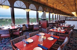 Un restaurante con varias mesas y sillas, amplias ventanas que dan al océano y lámparas colgantes.